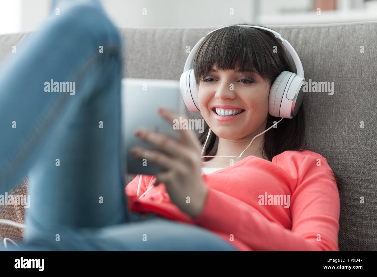 Lächelnde junge Frau zu Hause auf der Couch entspannen, trägt Kopfhörer, sie mit Hilfe einer digitalen-Tablets, ansehen von Videos und Musik online hören Stockfoto