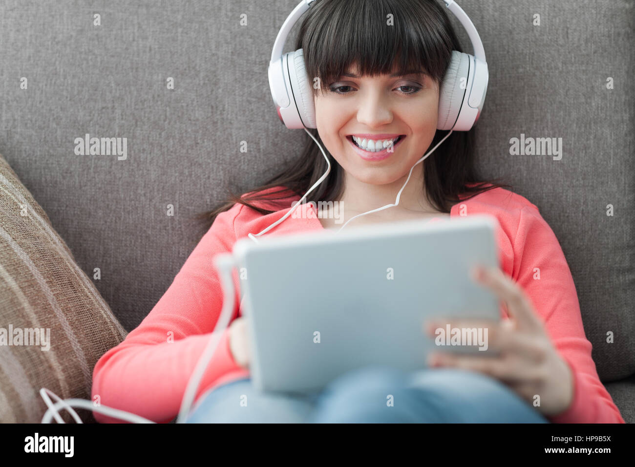 Lächelnde junge Frau zu Hause auf der Couch entspannen, trägt Kopfhörer, sie mit einem digital-Tablette und Teilnahme an Online-Kursen Stockfoto