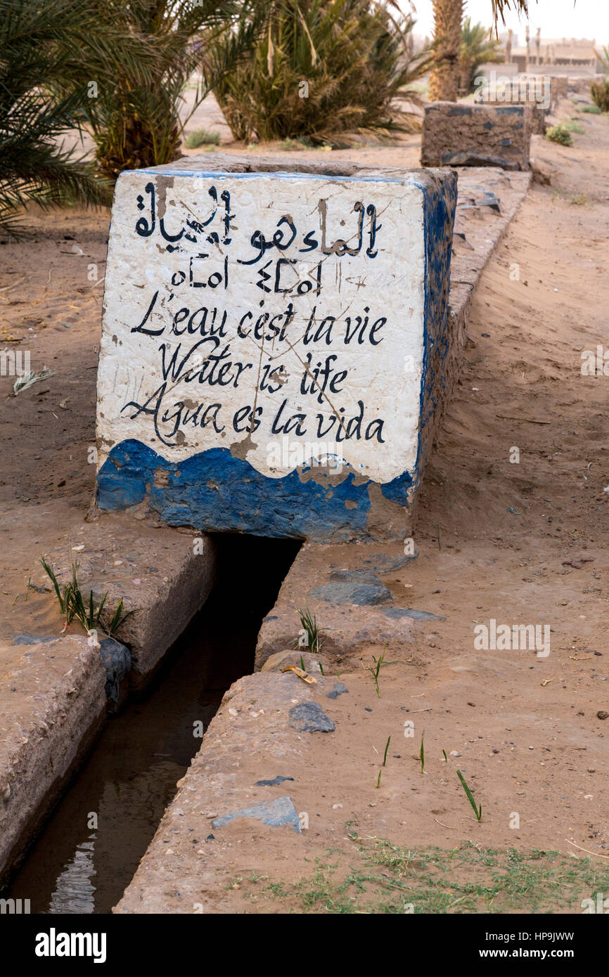 Merzouga, Marokko.  Bewässerungskanal Wassertragen Landwirte Plots in der Oase Merzouga.  "Wasser ist Leben" in fünf Sprachen. Stockfoto