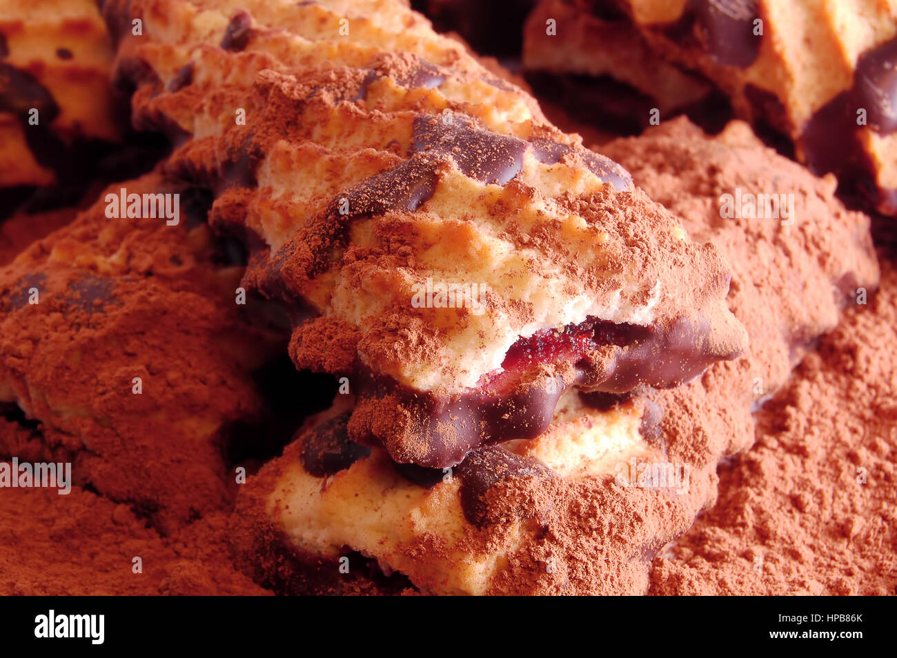 Haufen von Spritzgebäck mit Marmelade und Schokoladenglasur auf die verstreuten Kakaopulver hautnah. Stockfoto