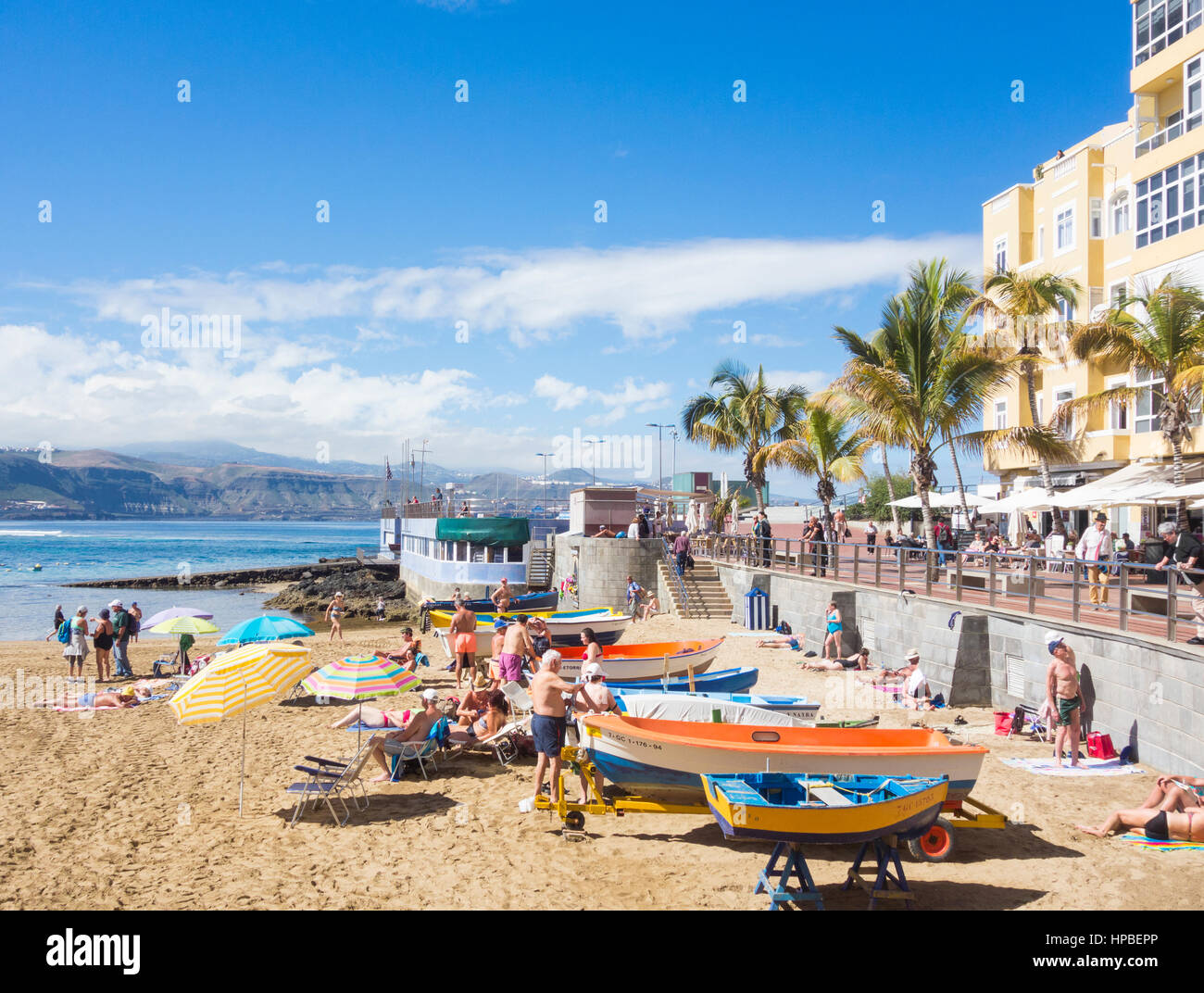 Menschen, die zum Sonnenbaden in der Nähe von bunten Fischerbooten am La Puntilla am Strand von Las Canteras in Las Palmas, Gran Canaria, Kanarische Inseln, Spanien Stockfoto