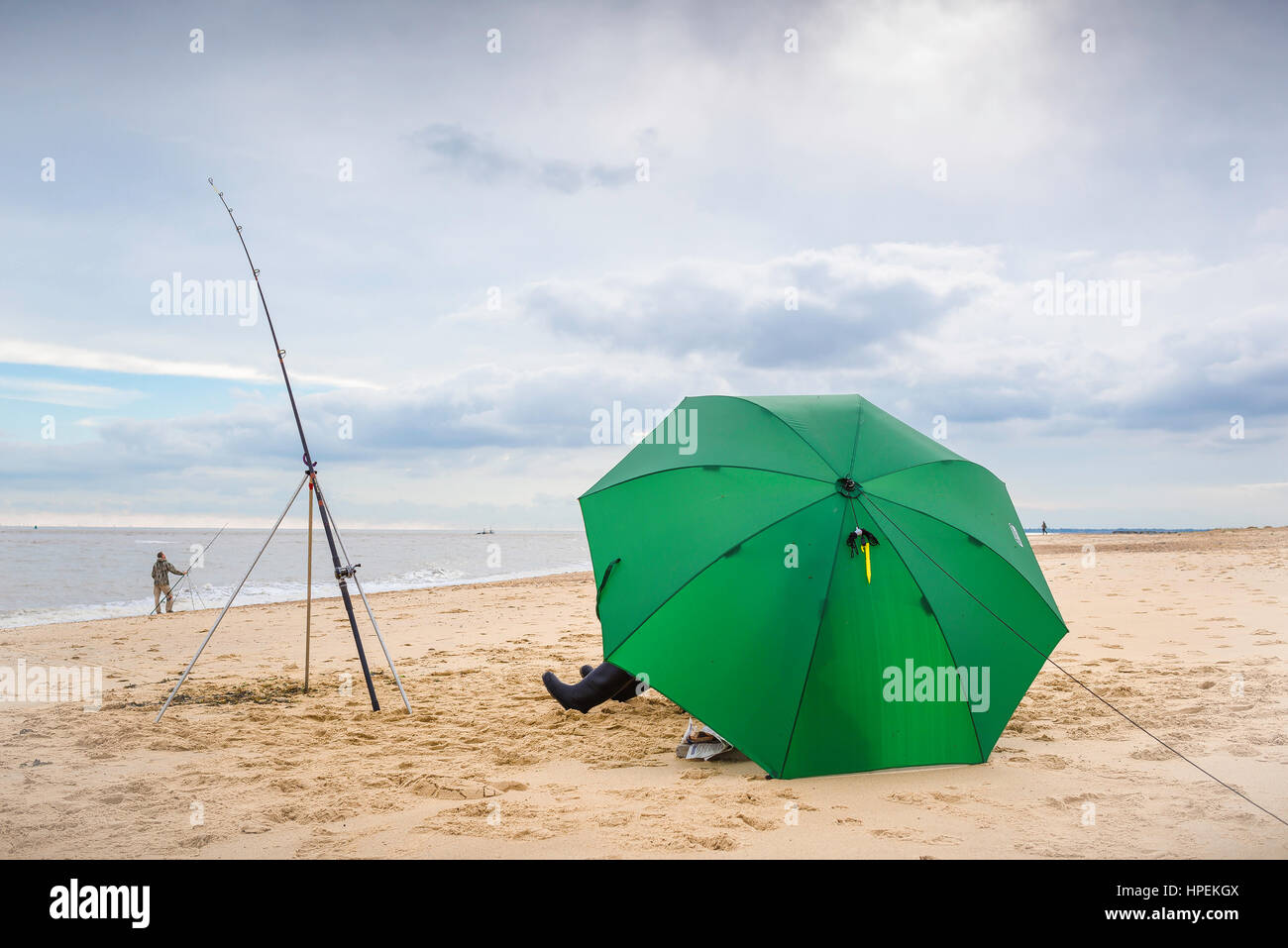 Angeln an der britischen Küste, Blick auf einen Fischer, der von einem großen Regenschirm abgeschirmt wird und geduldig darauf wartet, dass ein Fisch beißt, Felixstowe, Suffolk Coast, England, Großbritannien. Stockfoto