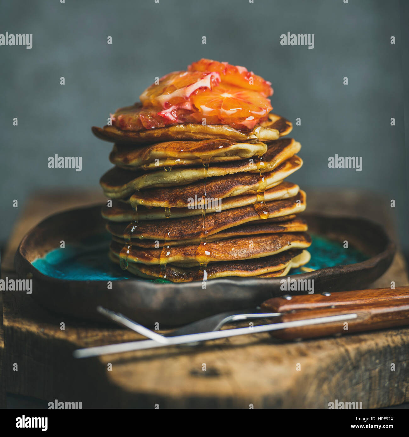 Hausgemachte Pfannkuchen mit Honig, blutigen Orangenscheiben und Minze zum Frühstück auf blaue Keramik-Platte über rustikalen Holzbrett, Sperrholz-Wand in grau Stockfoto
