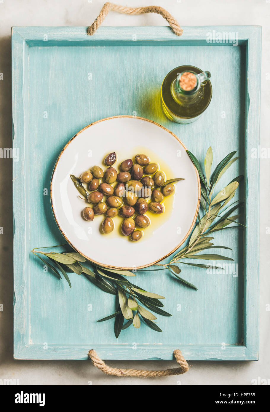 Eingelegte grüne mediterrane Oliven auf weißen Keramikplatte, Olivenbaum Zweig und natives Olivenöl in Glasflasche in hell blau gemalte Holztablett ov Stockfoto