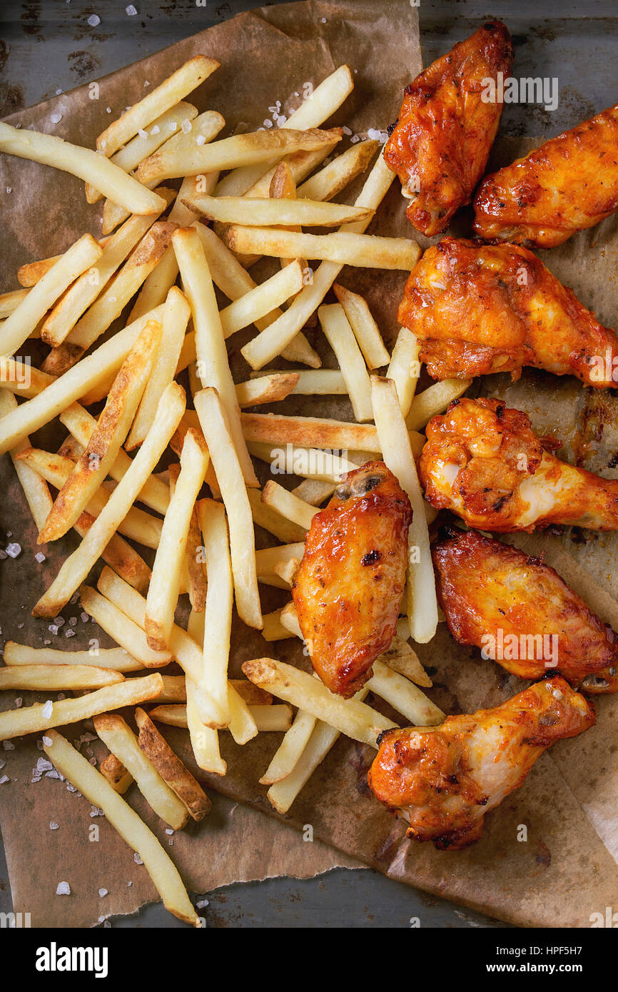 Fast-Food Bratkartoffeln würziges Huhn Beine, Flügel und Pommes mit Salz auf Backpapier über alten rostigen Metall Hintergrund serviert. Ansicht von oben Stockfoto