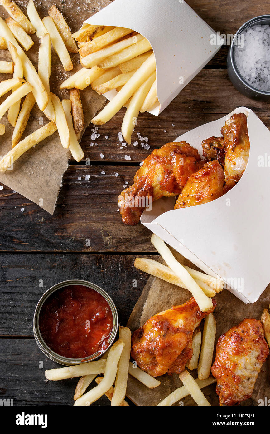 Fast-Food gebraten Huhn Beine, Flügel und Pommes frites Kartoffeln in Lunch-Boxen mit Salz und Ketchup serviert auf Backpapier über alte dunkle wo Soße Stockfoto
