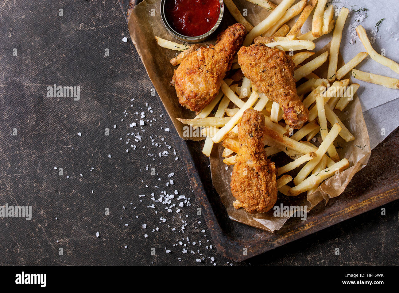 Fast-Food Bratkartoffeln knusprige Chicken Beine und Pommes mit Salz und Ketchup-Sauce, serviert auf Backpapier in alten rostigen Backblech über dunkle text Stockfoto