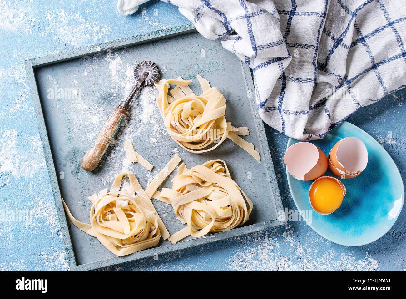 Frisches rohes hausgemachte verdrehten Teigwaren Tagliatelle mit Eigelb und Pasta Eierschneider Holztablett mit Küchentuch über leichte blaue konkrete backgr Stockfoto