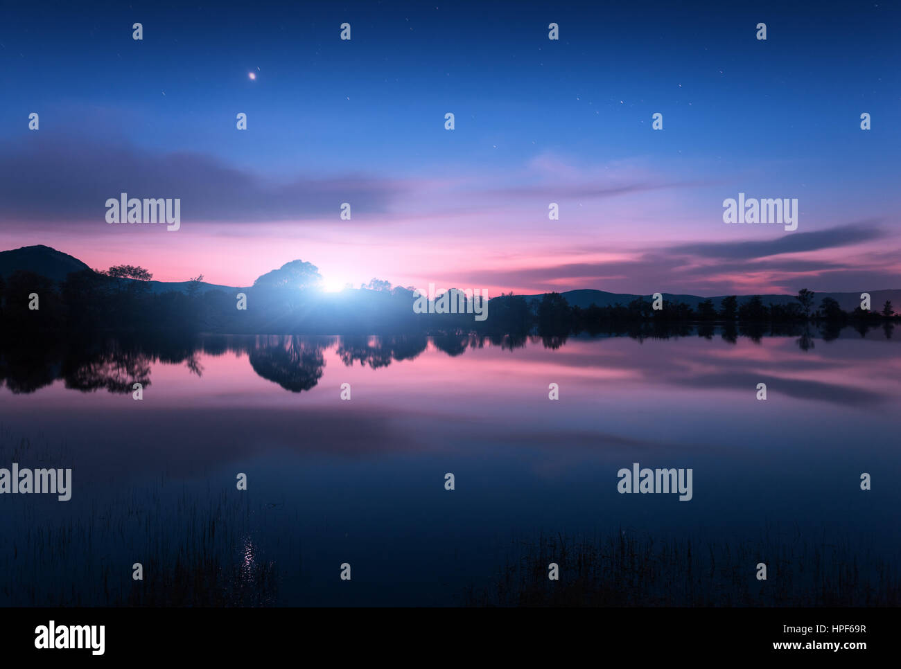 Bergsee mit Mondaufgang in der Nacht. Nacht-Landschaft mit Fluss, Bäume, Hügel, Mond und bunten blauer Himmel mit Wolken spiegelt sich im Wasser in Dämmerung Stockfoto