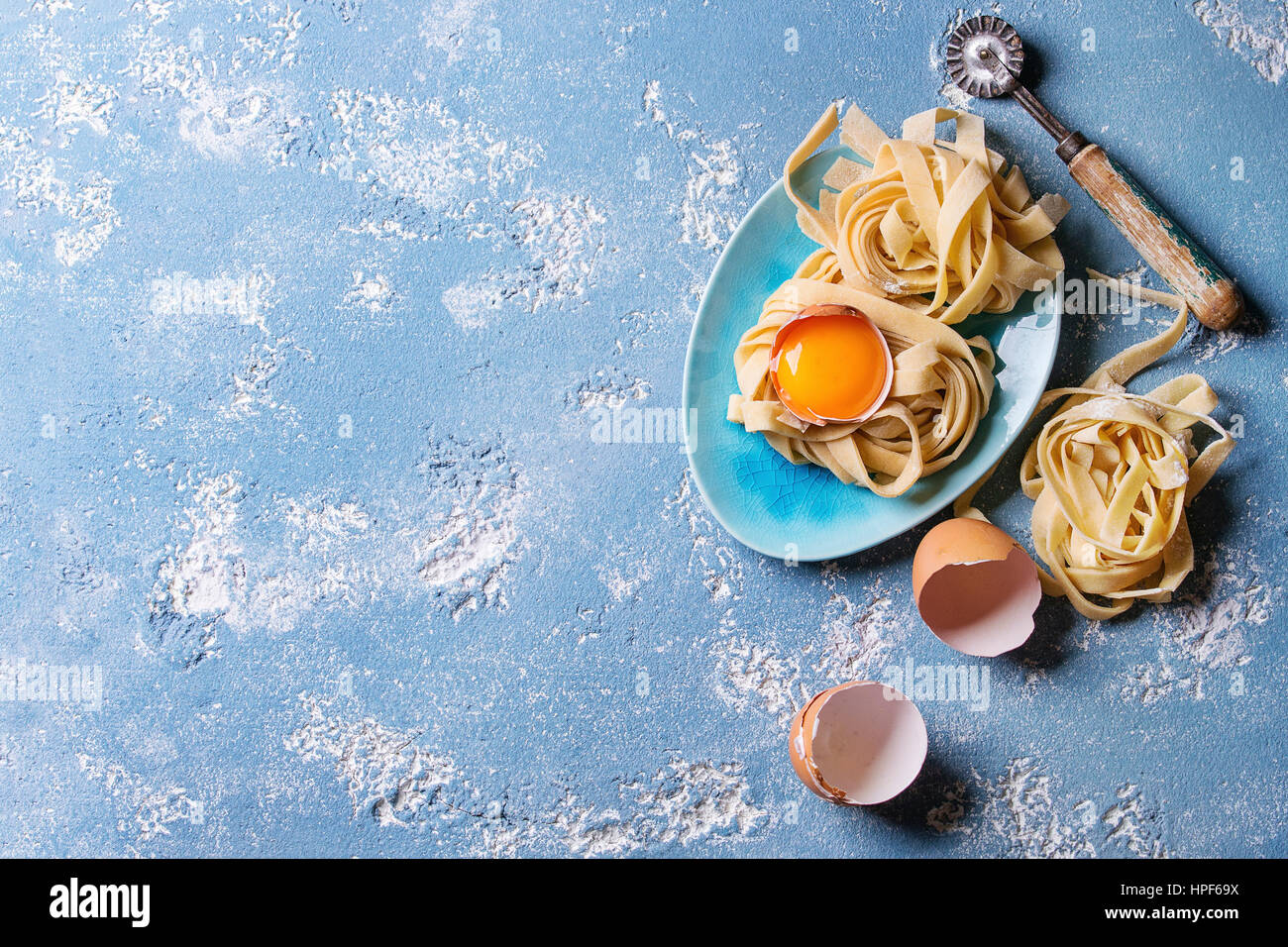 Frisches rohes hausgemachte verdrehten Teigwaren Tagliatelle mit Eigelb, Schale und Pasta Eierschneider Holztablett mit Küchentuch über blaue Leichtbeton Stockfoto