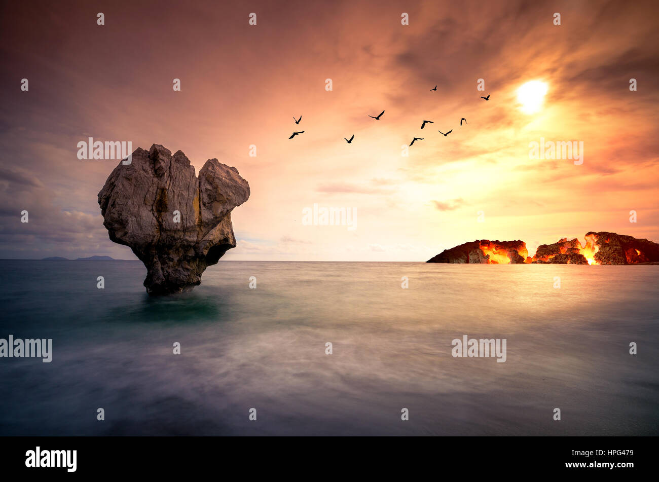 Fine Art mit Lonely Rock Skulptur im Meer mit Silhouetten von fliegenden Vögeln und eine brennende Insel unter den Sonnenuntergang. Stockfoto