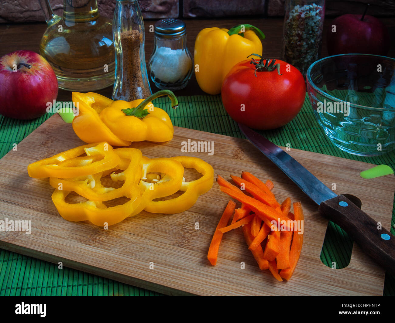 Große gelbe saftige Scheiben Paprika Ringe auf einem Holzbrett auf einem Küchentisch mit Karotten, rote Tomaten, Äpfel, Pflanzenöl, Salz, Gewürze Stockfoto