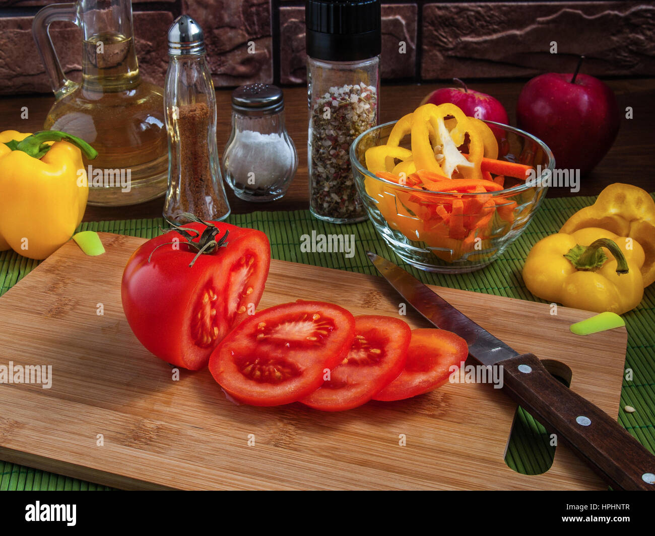 Große gelbe saftige Scheiben Paprika Ringe auf einem Holzbrett auf einem Küchentisch mit Karotten, rote Tomaten, Äpfel, Pflanzenöl, Salz, Gewürze Stockfoto