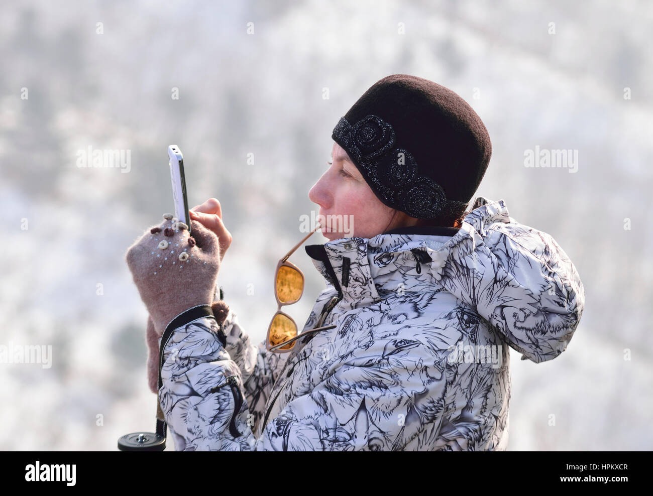 Junge Frau im Winter Kleidung stehen eine Nachricht auf dem Handy zu schreiben hält Gläser in Mund Stockfoto