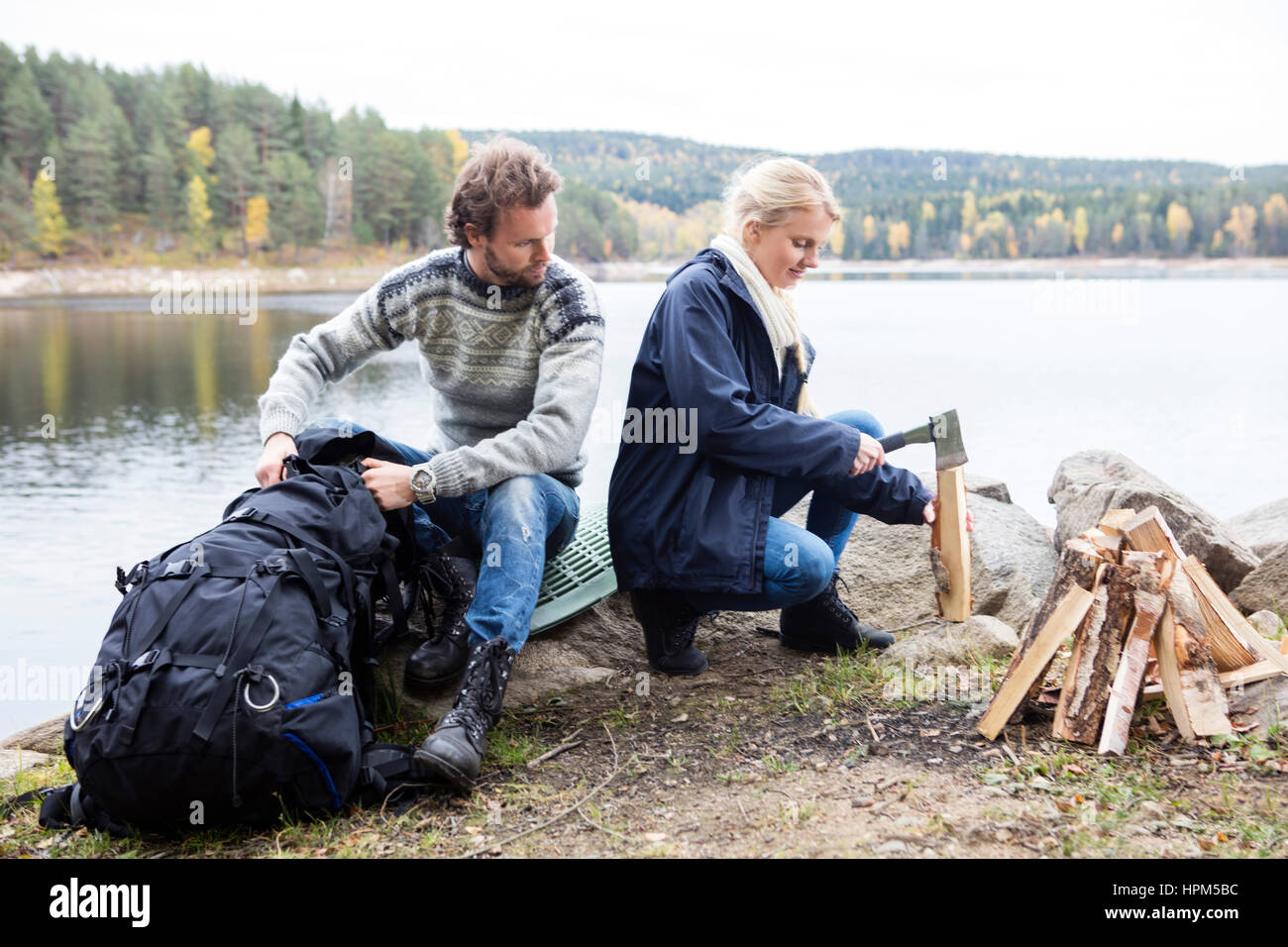 Junger Mann mit Blick auf Frau Schneiden von Holz am Seeufer bei camping Rucksack Stockfoto