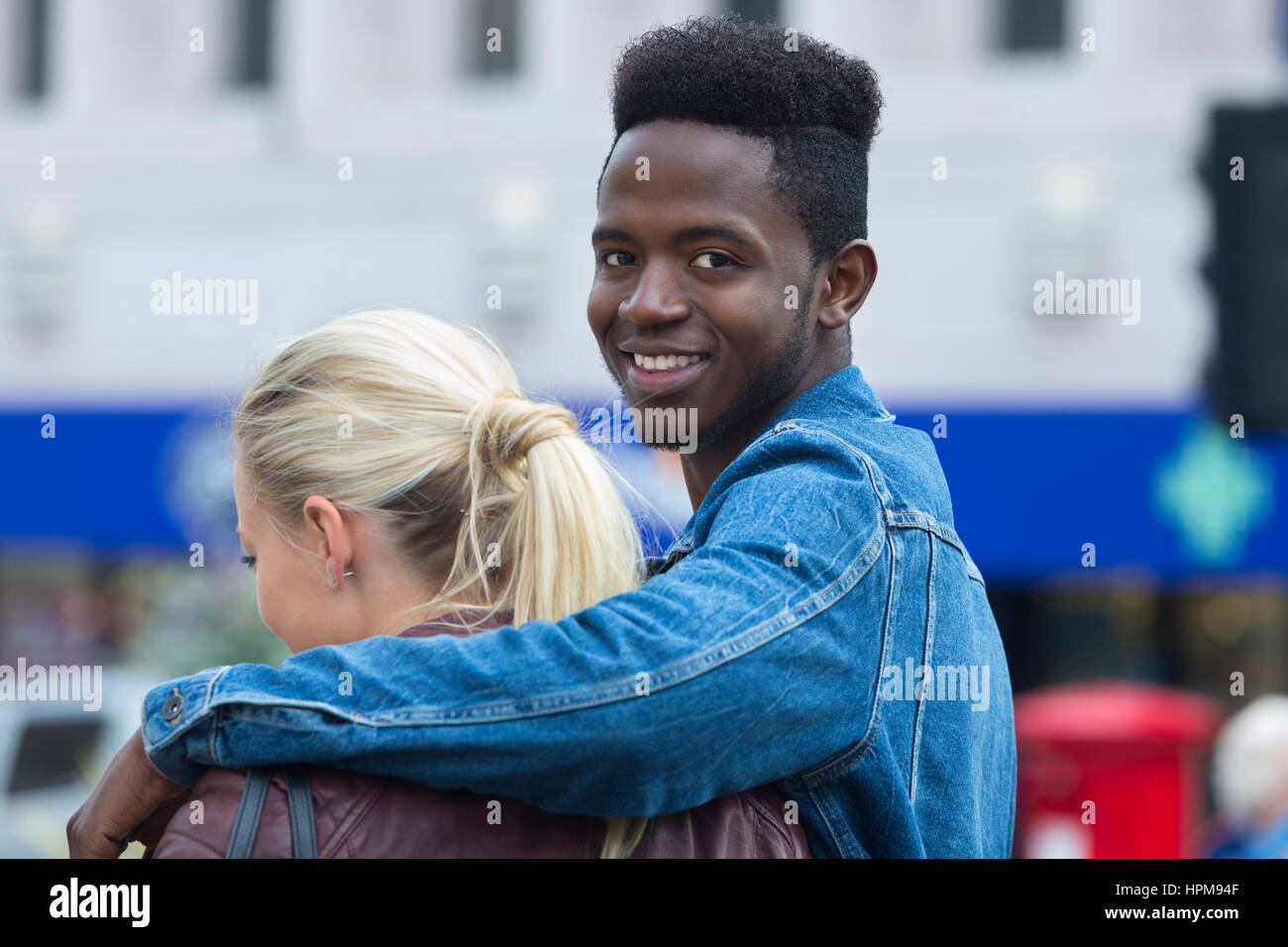 Ein glücklicher junge Mann lächelt in die Kamera, als er seinen Arm um seine Freundin Schulter umschließt. Sie hat ihre Rücken zur Kamera. Stockfoto