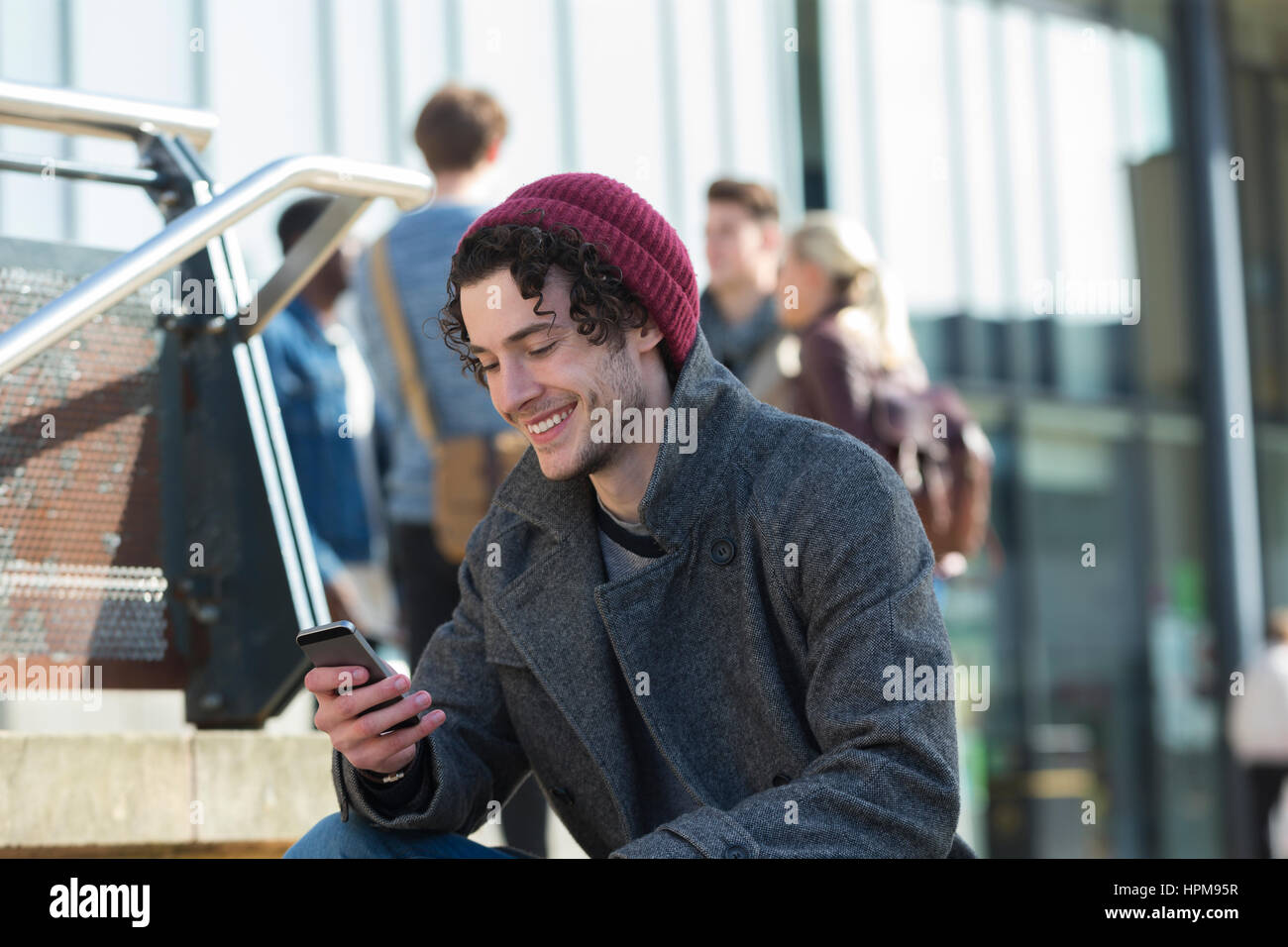 Ein glücklicher junge Mann lächelt, als er sein Handy in der Stadt verwendet. Er steckt sich Warm für das kalte Wetter und trägt einen Hut. Stockfoto
