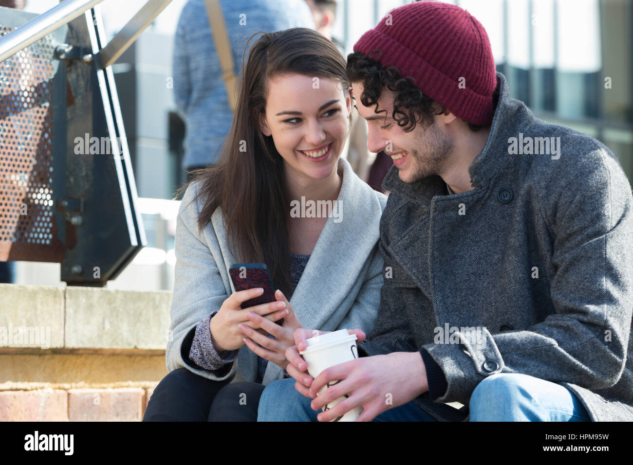 Eine Nahaufnahme von ein glückliches junges Paar Lächeln, als sie auf einige sitzen Schritte in der Stadt mit Blick auf ein Mobiltelefon. Das Männchen hält ein heißes Getränk in ein Stockfoto