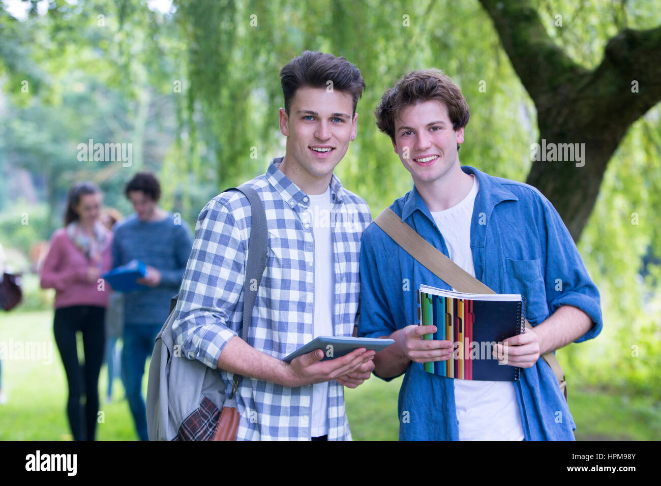 Zwei glückliche männliche Studenten Lächeln, als sie im Freien und Studie zu entspannen. Sie halten ihre Taschen, Bücher und ein digital-Tablette, wie sie in die Kamera blickt. Stockfoto