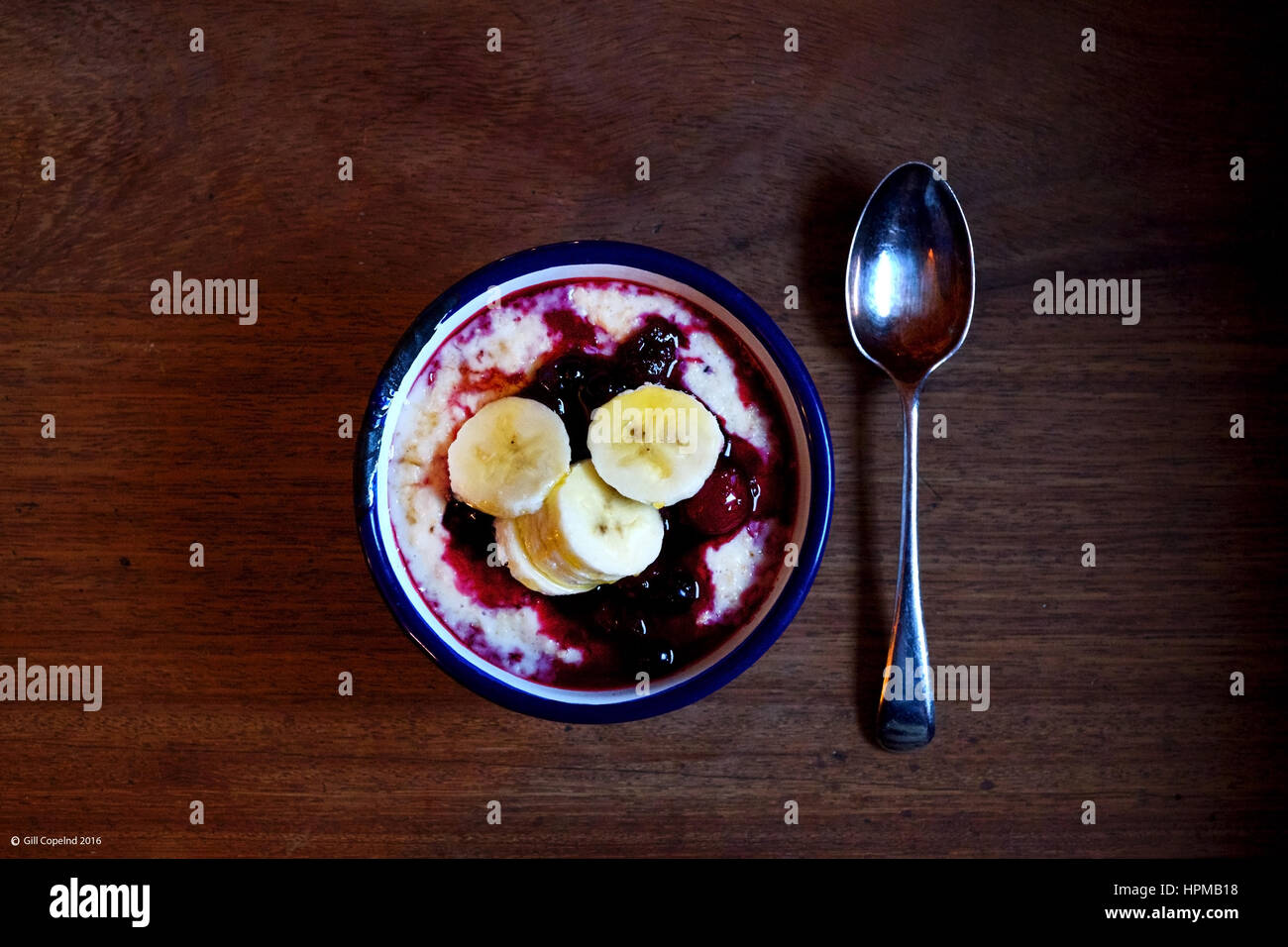 eine Schüssel Brei mit rote Himbeer Frucht und Banane in ein weißes Metall Schüssel mit einem blauen Rand einer einzigen Silberlöffel daneben auf einem Holztisch, t Stockfoto