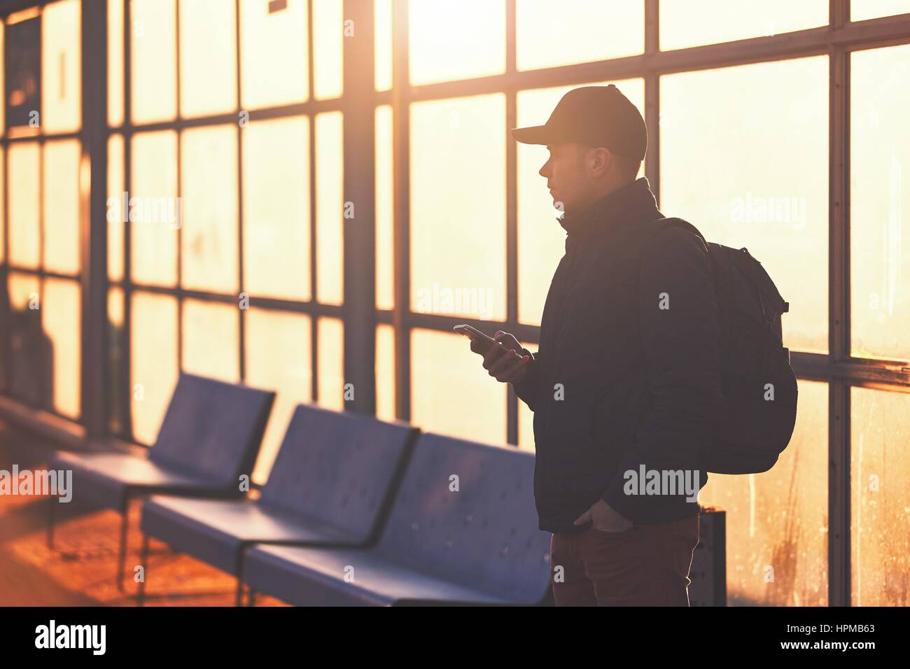 Junge Reisende mit Handy in der hand warten an der Bushaltestelle während des Sonnenuntergangs. Stockfoto