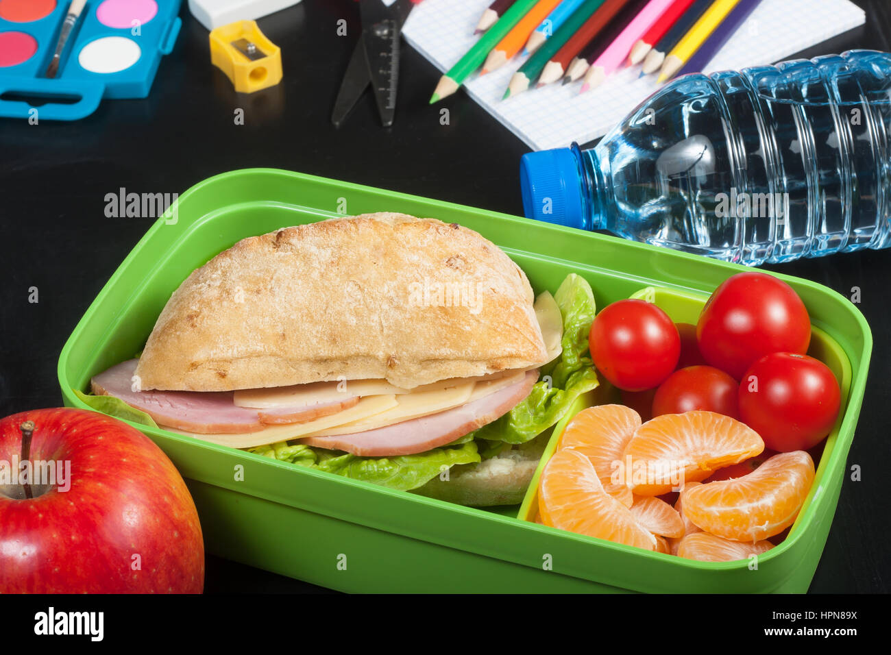 Schulspeisung. Sandwich, kleine Tomaten, Mandarine, Apfel in Kunststoff Lunch-Box und eine Flasche Wasser an schwarze Tafel. Stockfoto