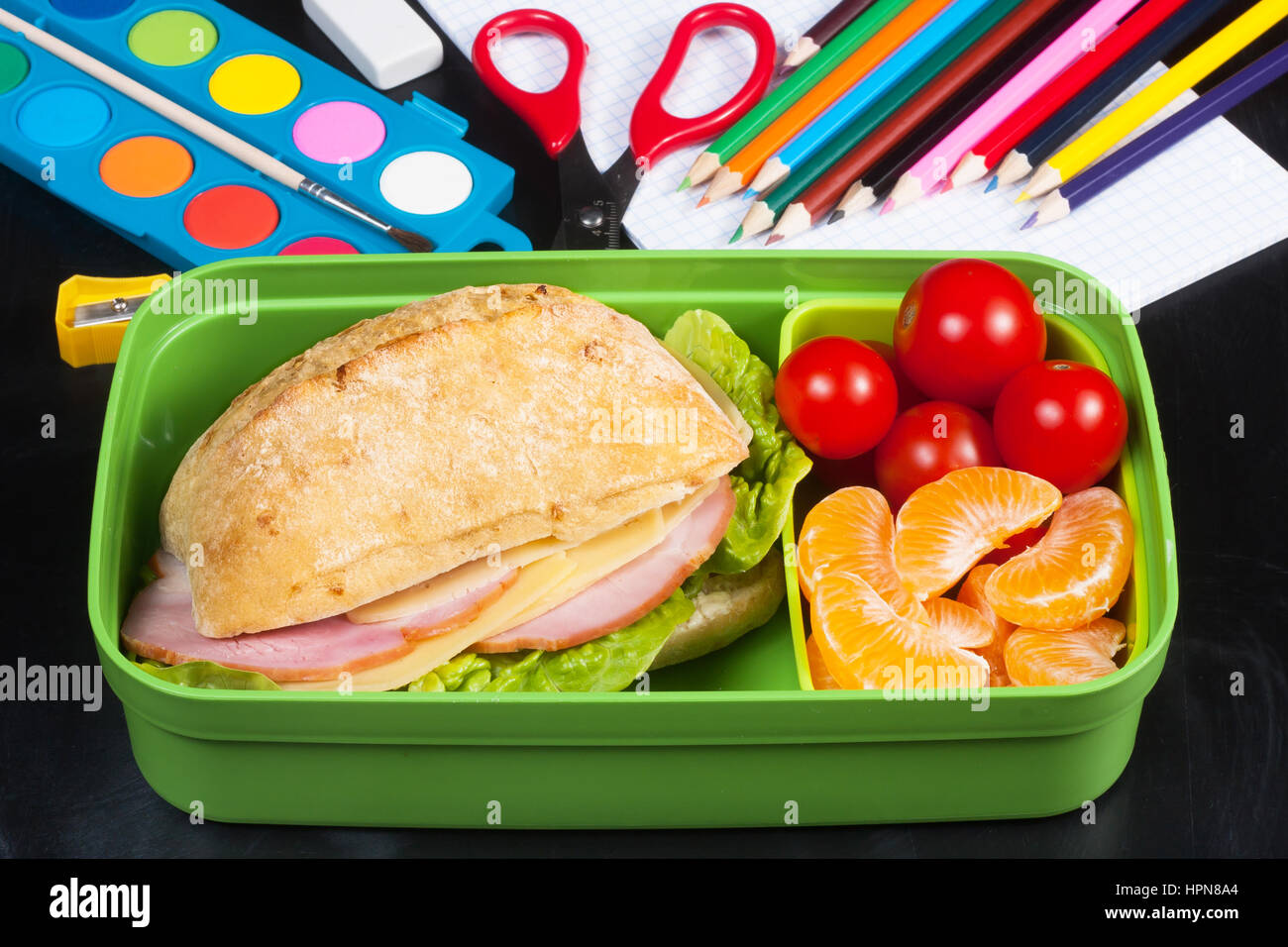 Schulspeisung. Sandwich, kleine Tomaten, Mandarine in Kunststoff Lunchpaket an schwarzen Tafel. Stockfoto