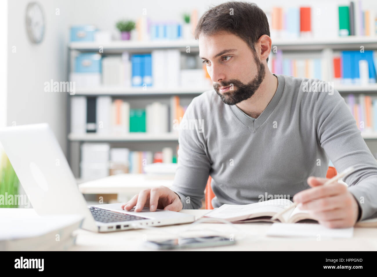 Junger hübscher Mann am Schreibtisch sitzen, ein Buch und Verbindung zum Internet mit einem laptop Stockfoto