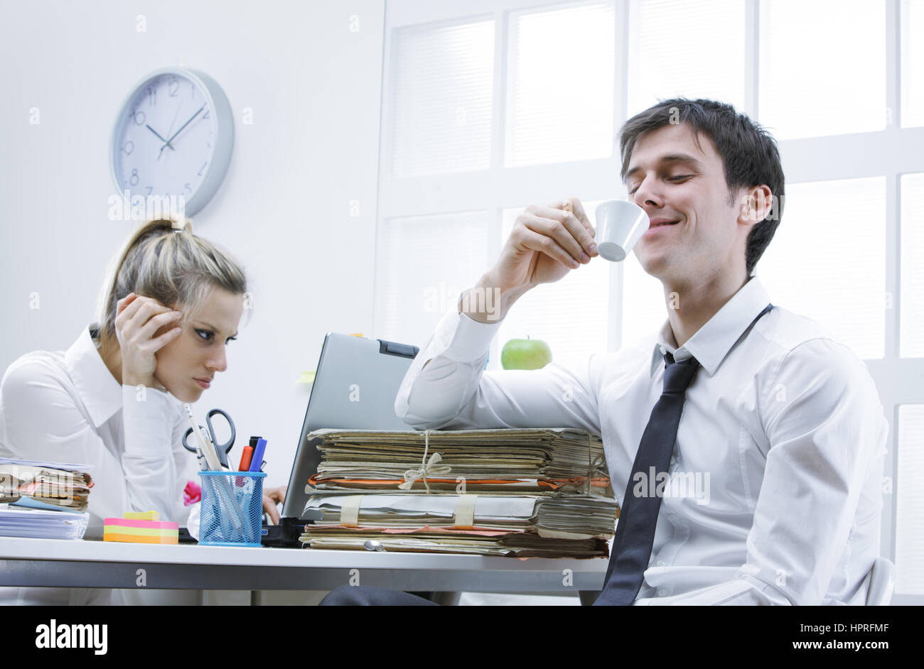 Business-Mann genießt eine heiße Tasse Kaffee im Büro, verärgert Kollegin im Hintergrund Stockfoto