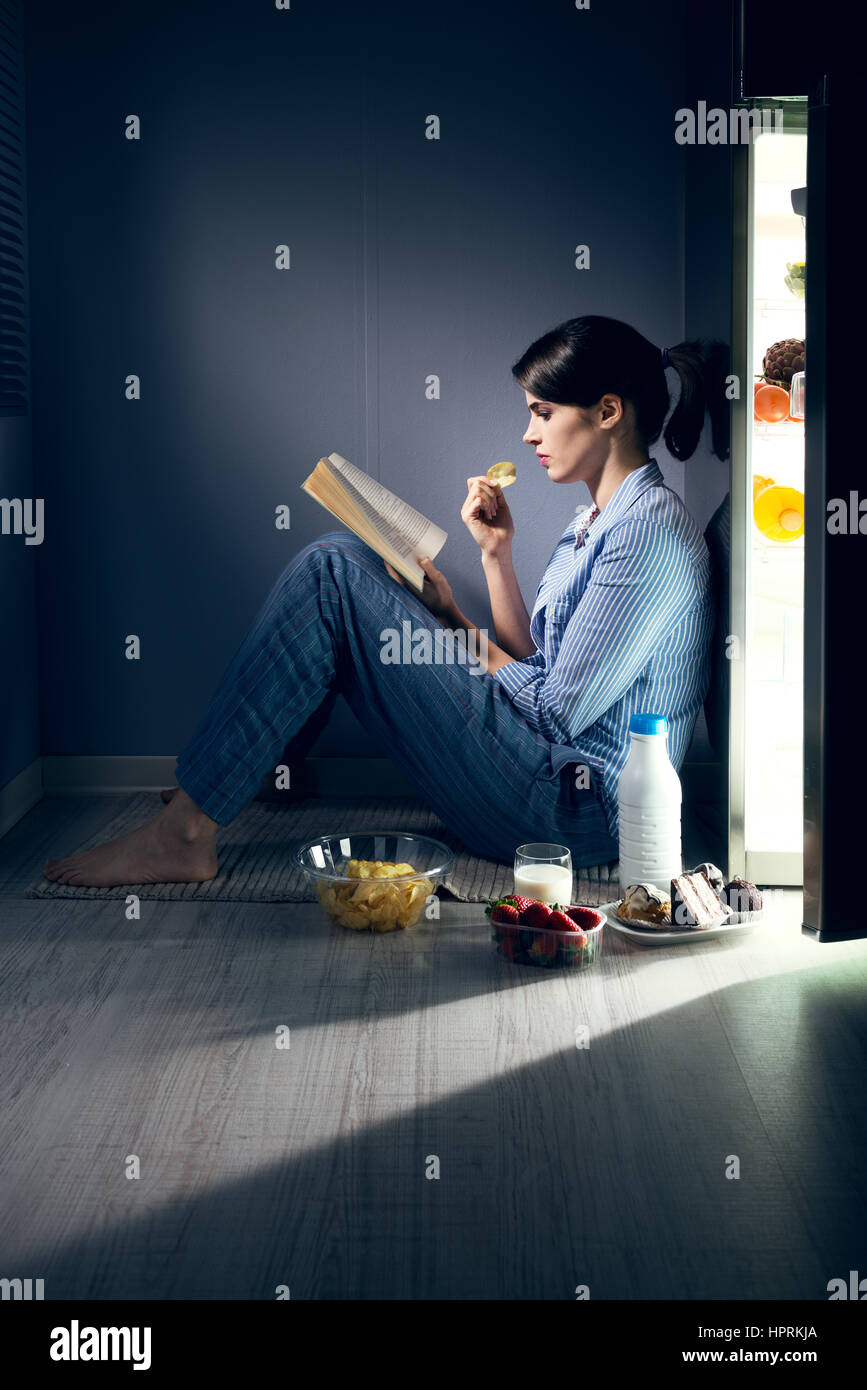 Schlaflose Frau sitzt auf dem Küchenboden ein Buch lesen und Essen. Stockfoto