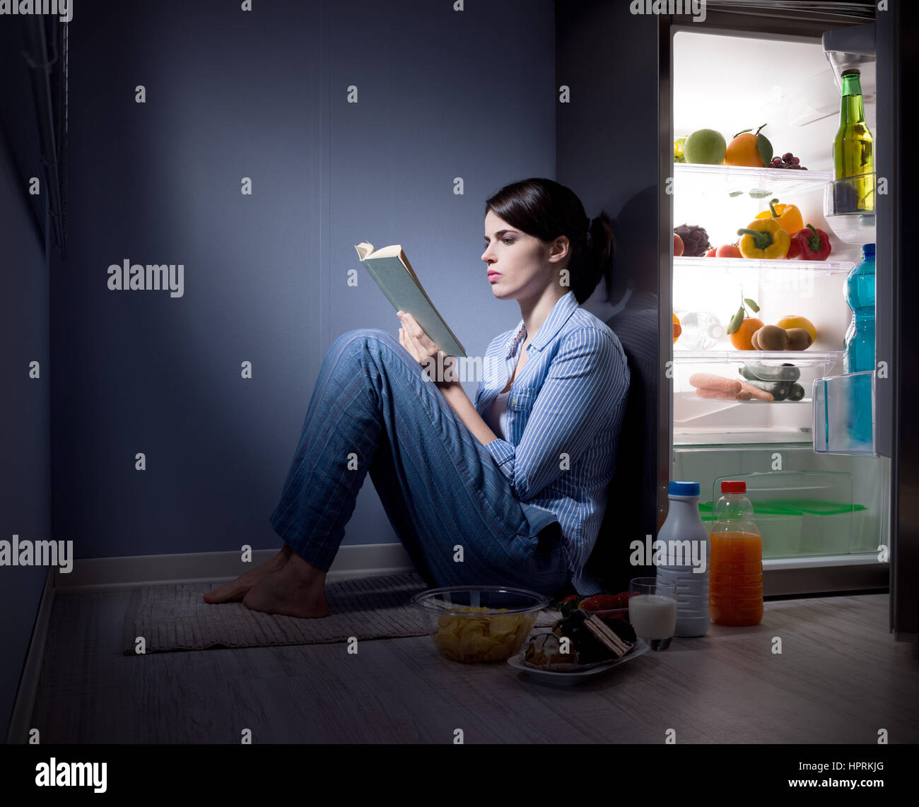 Schlaflose Frau sitzt auf dem Küchenboden ein Buch lesen und Essen. Stockfoto