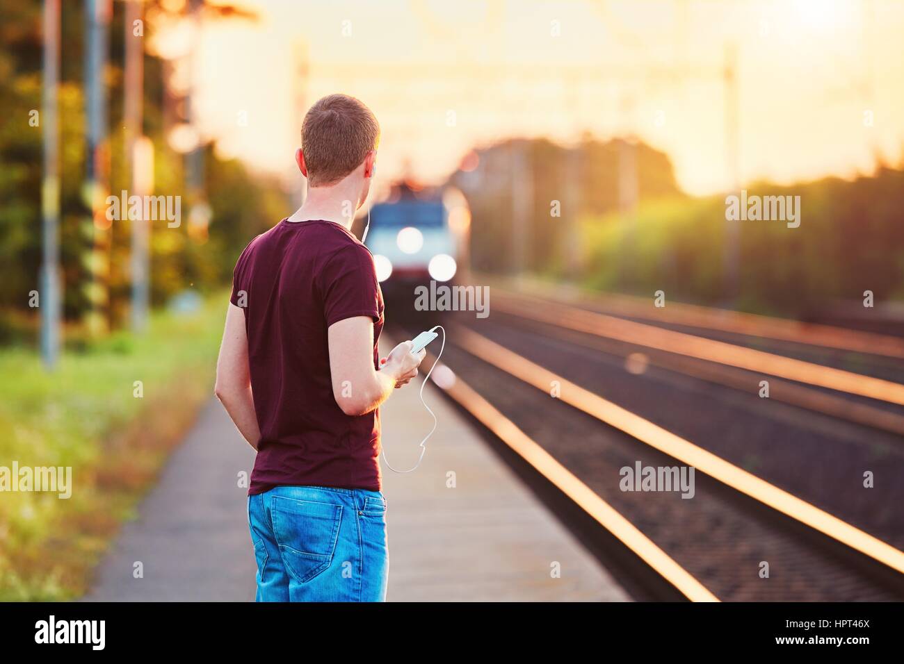 Bahnhof in den Sonnenuntergang. Junger Mann mit Smartphone in der Hand wartet auf den Zug. Stockfoto