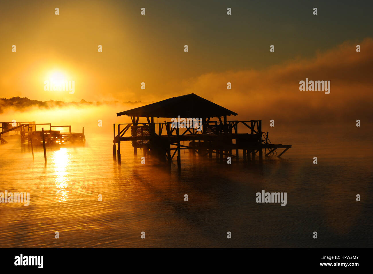 Am frühen Morgennebel steigt vom See Chicot Lake Village, Arkansas.  Dock und Boot Holzhaus sind Silhouette. Goldenes Licht deckt See. Stockfoto