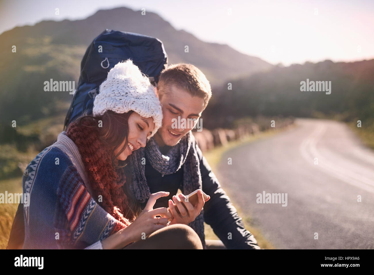 Junges Paar mit Rucksack wandern, ausruhen und mit Handy auf sonnigen, abgelegenen Straße Stockfoto