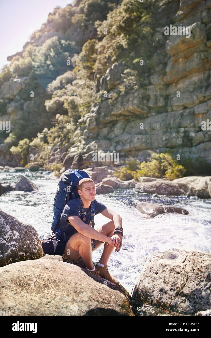 Junger Mann mit Rucksack wandern, ruht auf Felsen am sonnigen Fluss Stockfoto