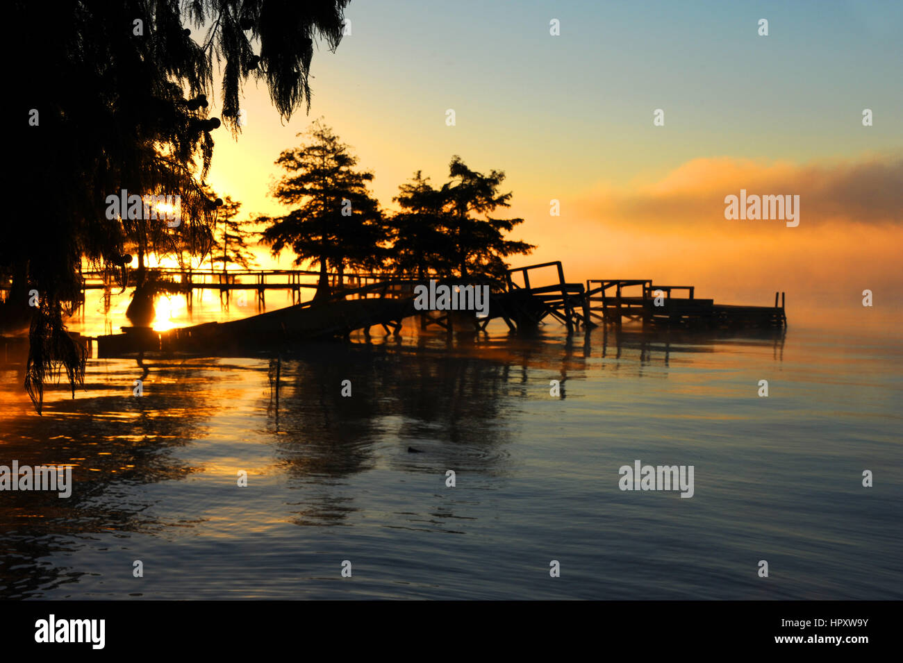 Morgennebel schmiegt sich einen verwesenden Pier am See Chicot im östlichen Arkansas.  Sunrise-Tönungen Wasser Orange und gelb. Stockfoto