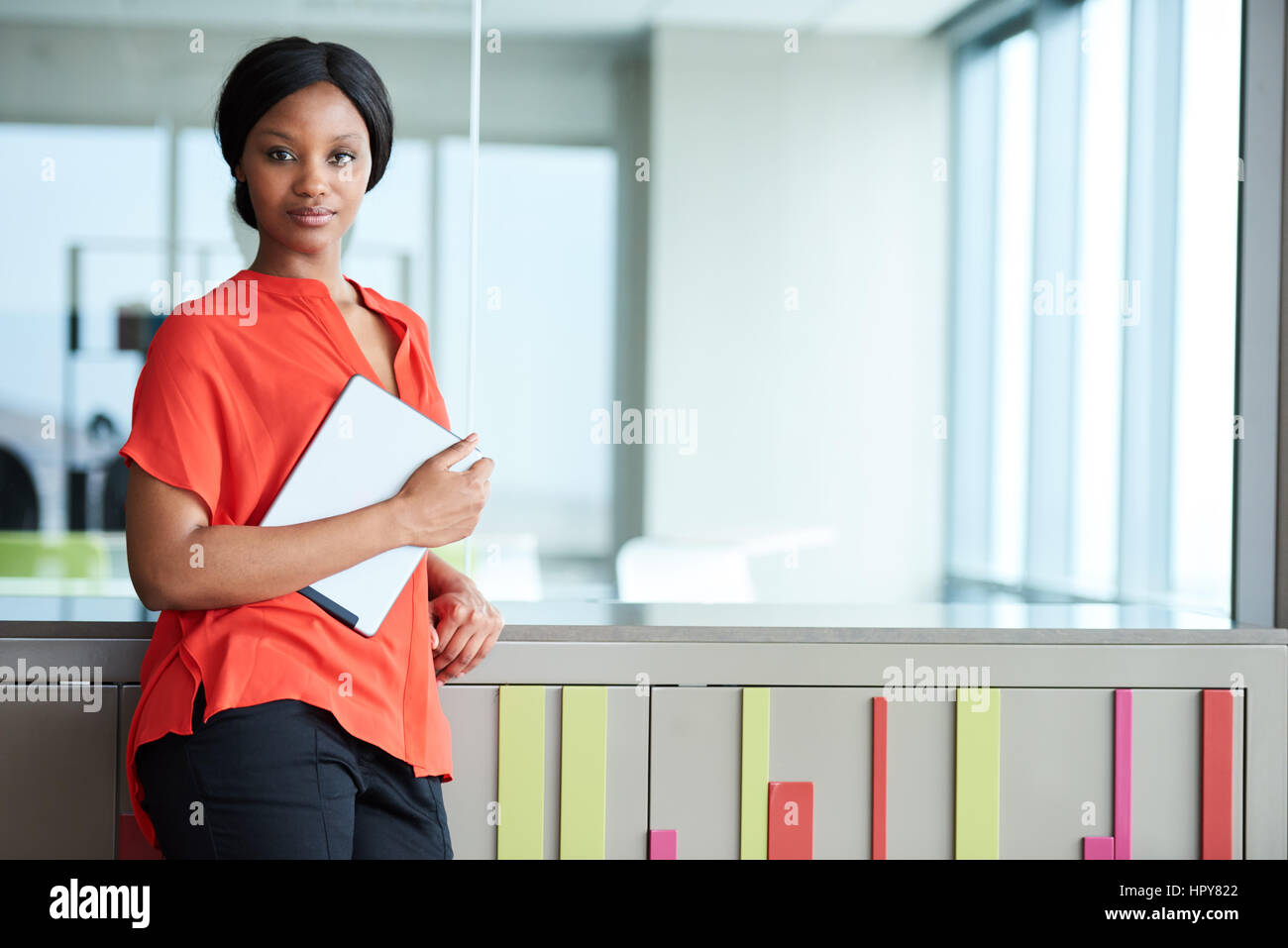 Geschäftsfrau trägt eine helle orange Bluse und hält eine digitale-Tablette während stehend in einem bunten Geschäftsumfeld, Blick geradeaus in th Stockfoto