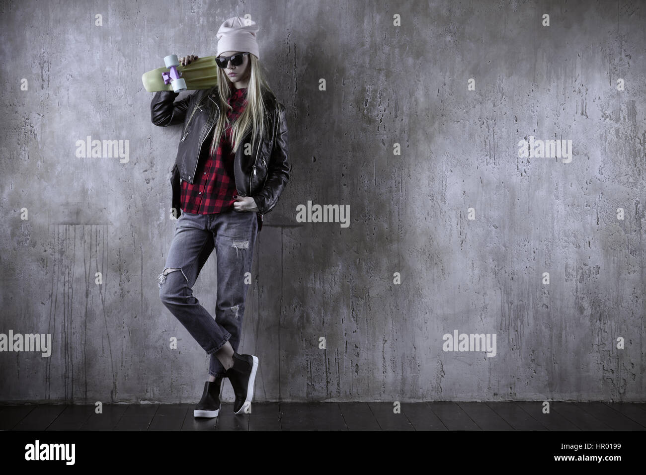 Blonde Teenager Hipster Mädchen in schwarzen Jacke und schwarze Sonnenbrille und Hut mit blauen Skateboard. Vor Betonwand. Stockfoto