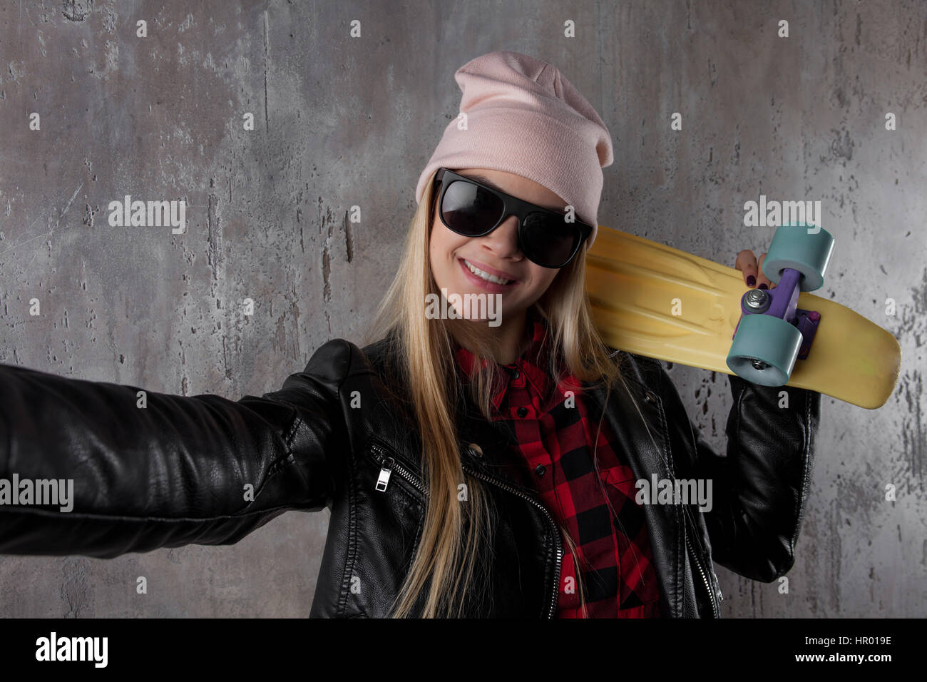 Blonde Teenager Hipster Mädchen in schwarzen Jacke und schwarze Sonnenbrille und Hut mit gelben Skateboard machen Selfie. Vor Betonwand. Stockfoto