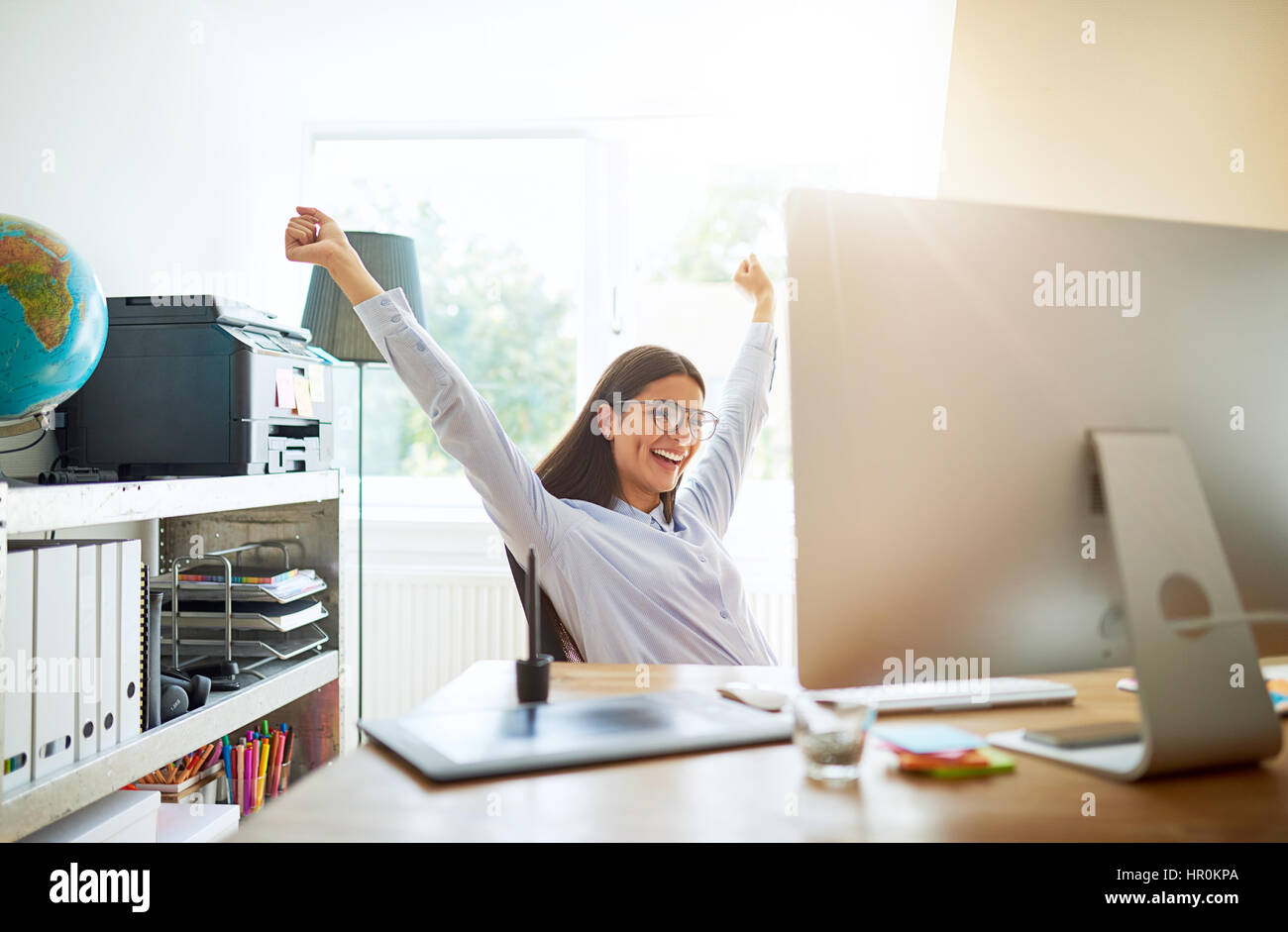 Einzelne Jugendliche begeistert Frau feiern etwas mit beiden Armen nach oben verlängert, sitzend am Schreibtisch im kleinen Büro Stockfoto