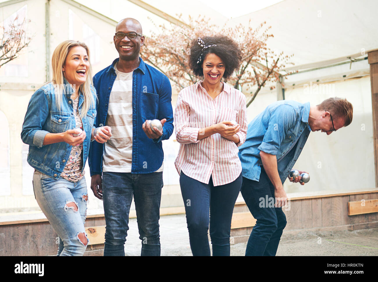Gruppe von vier Jugendlichen Petanque Spieler tragen legere Kleidung stehend halten Metall Kugeln in die Hände spielen und lachen, Stockfoto