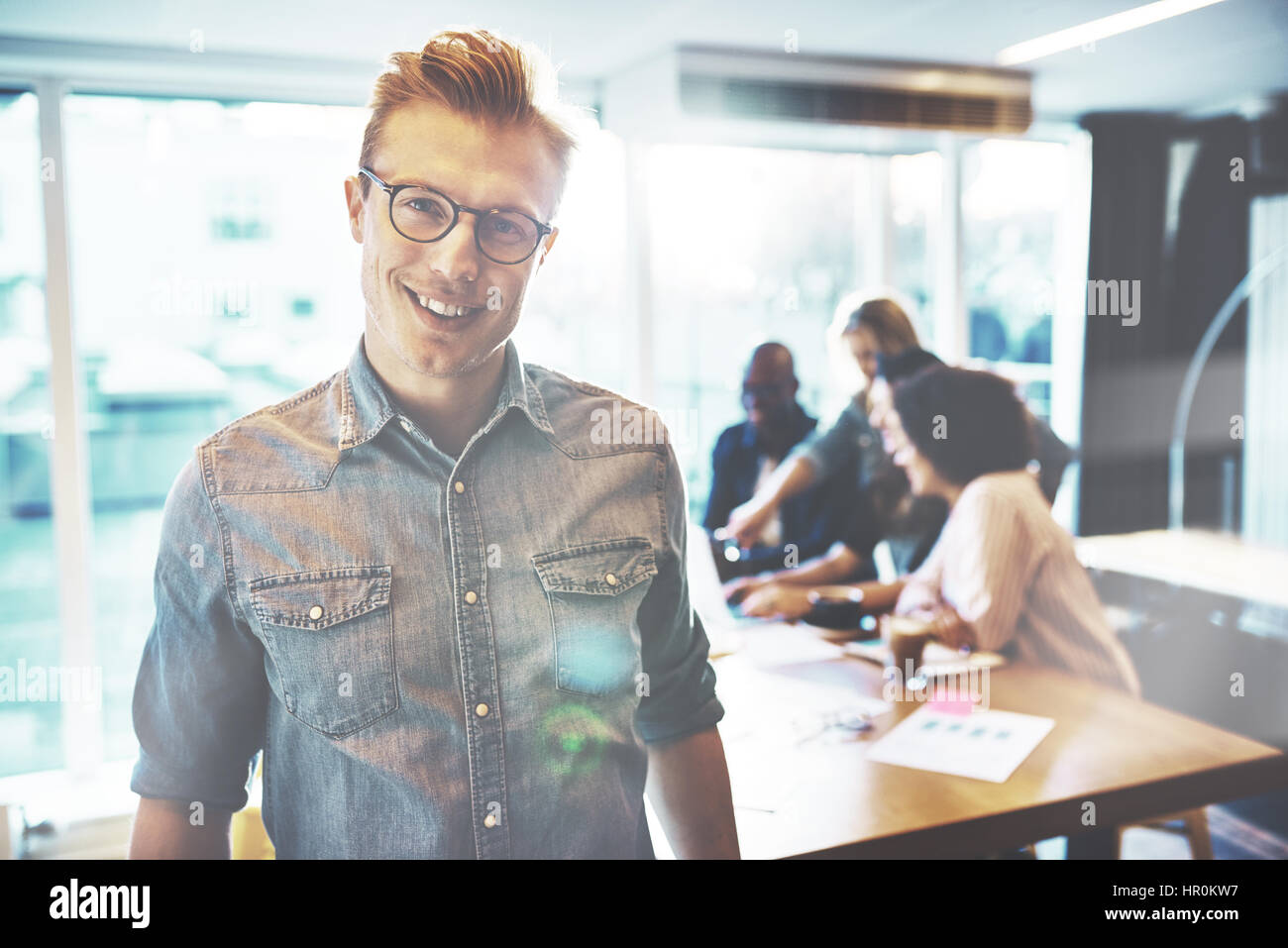 Schönen jungen Mann tragen Brillen und Freizeitkleidung Blick in die Kamera und lächelnd, in helles Büro mit Team arbeitet im Hintergrund stehend Stockfoto