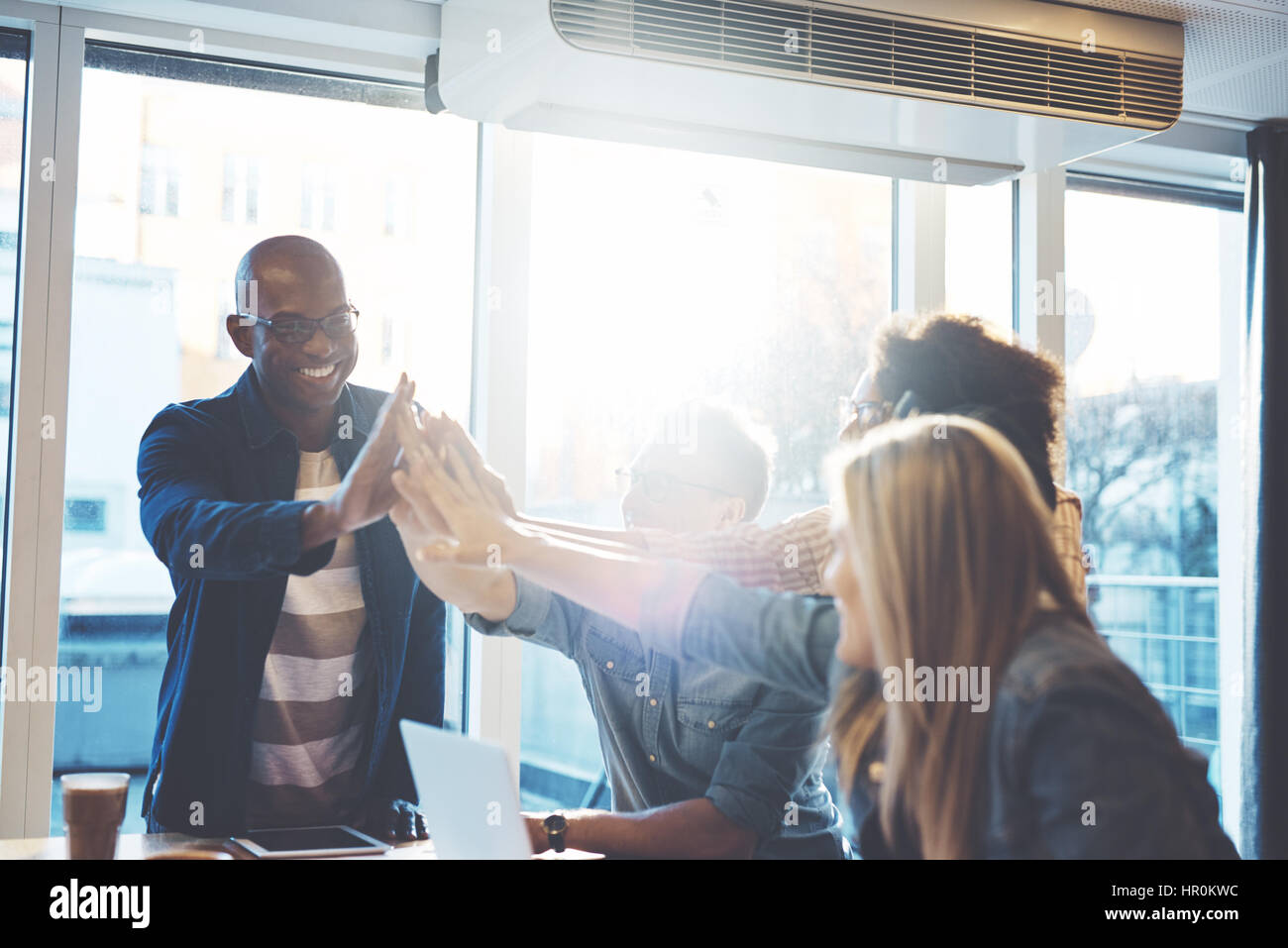 Junge Menschen in Freizeitkleidung gibt hohe Fives miteinander, als ob etwas gegen hellen Fenster im Café oder Business Office feiert Stockfoto