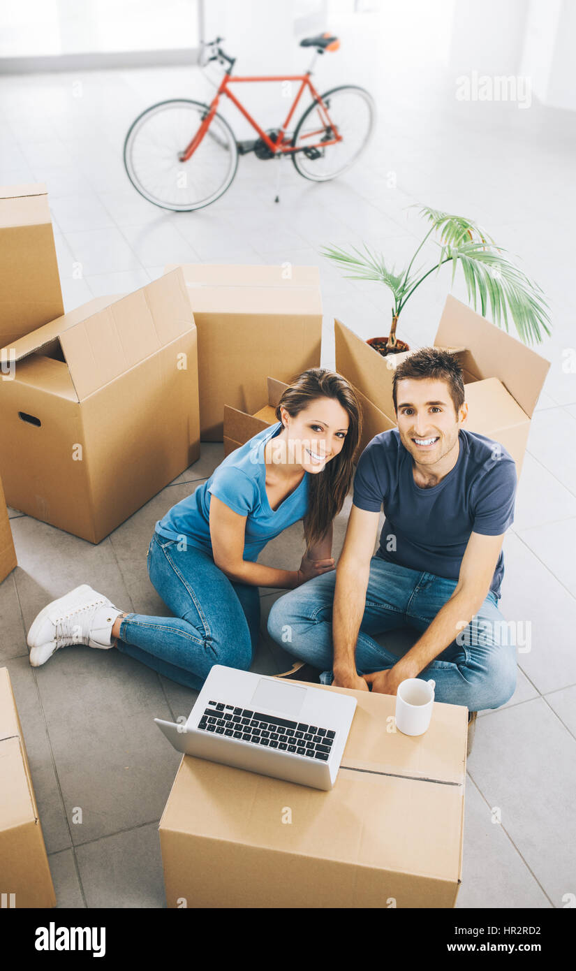 Glückliches junges Paar sitzt auf dem Boden ihres neuen Hauses, umgeben von Kartons, sind sie in die Kamera Lächeln. Stockfoto
