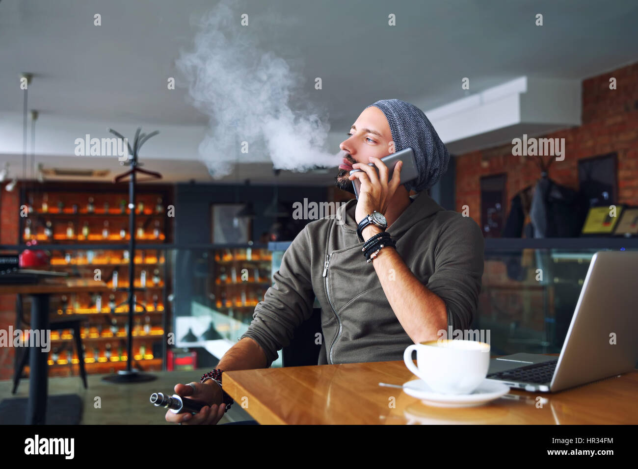 Junge hübsche Hipster Mann mit Bart sitzen im Café bei einer Tasse Kaffee, dampfen und Veröffentlichungen eine Wolke aus Dampf. Handy zu sprechen und haben eine kleine Pause. Stockfoto