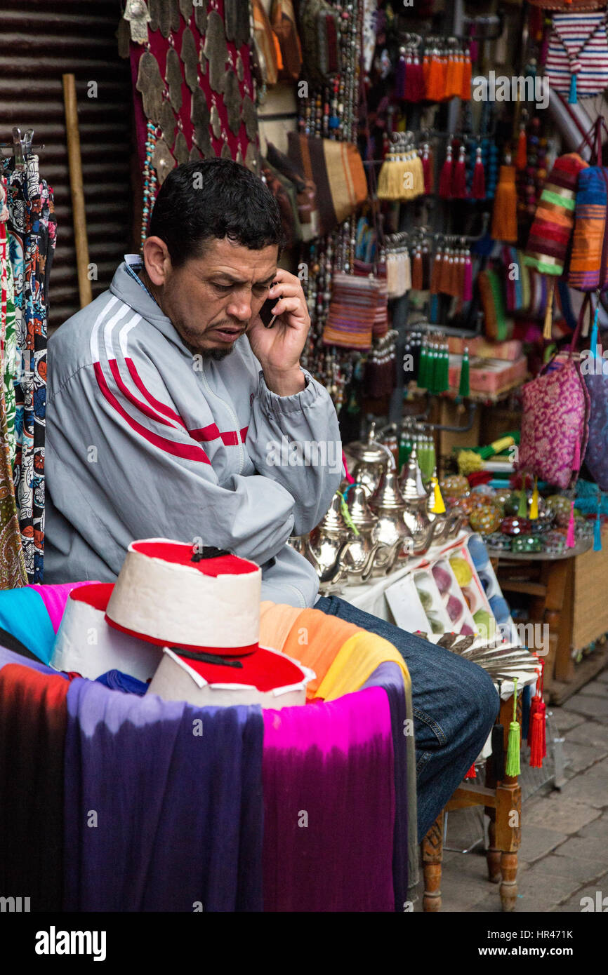Marrakesch, Marokko.  Shop-Anbieter von Fezen, Teekannen und dekorative Elemente hören auf sein Handy. Stockfoto
