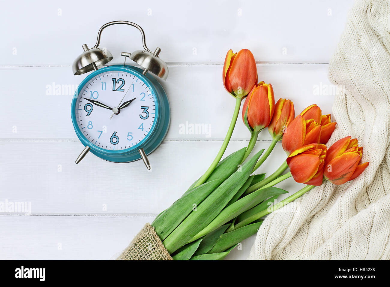 Wecker mit einem schönen Strauß Tulpen und eine kuschelige Decke, von oben in einem Flatlay Stil über eine hölzerne Tischplatte erschossen. Sommerzeit-Konz Stockfoto