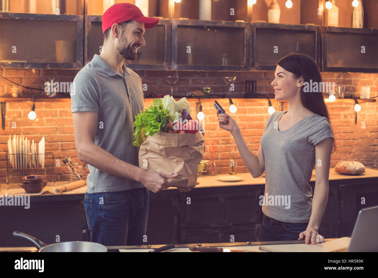 Lebensmittel-Lieferservice. Junger Mann bringt frischen Lebensmittel. Junge Frau mit Kreditkarte zu bezahlen. Schöne Loft Küche Interieur mit Glühbirnen Stockfoto