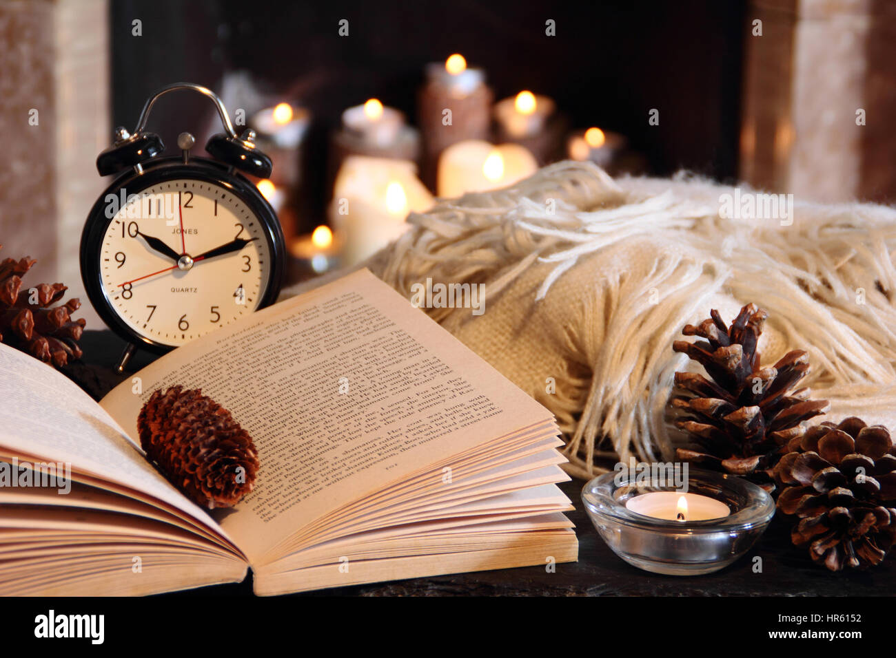 Lesen Sie ein Taschenbuch in der Nacht bei Kerzenschein neben einem offenen Kamin in einem gemütlichen englischen Haus mit einer Decke (werfen) im winter Stockfoto
