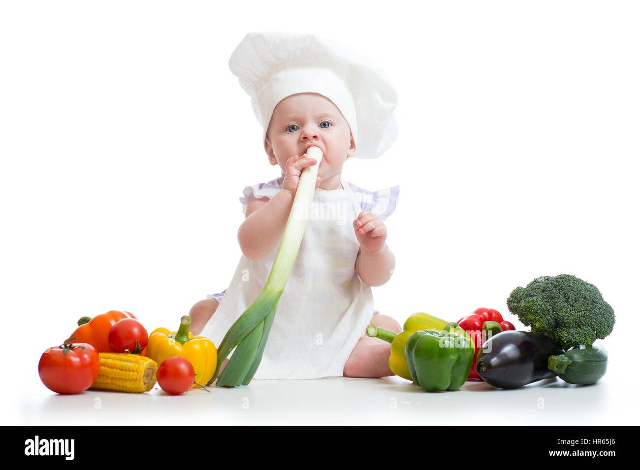 Kleines Mädchen, gekleidet in weißen Koch Hut und Schürze isst frische Zwiebel. Kind mit Gemüse: Popcorn, Brokolli, Paprika, Zucchini, Auberginen, Tomaten Stockfoto